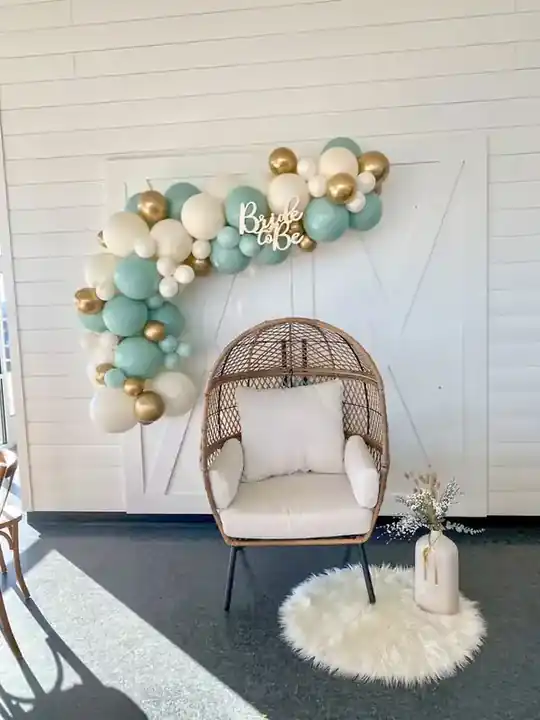 Teal, white and gold balloon garland curving over a wicker egg chair